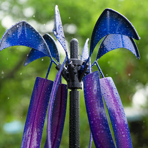 A close-up of a decorative metal garden sculpture with curved blue and purple leaf-like blades on a black central post, shown outdoors in the rain with water droplets and a blurred green background.