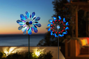This image captures the beauty of two windmills at dusk. The blue and red windmills stand out against the darkening sky and distant ocean. Lights illuminate the surrounding greenery, adding a warm and inviting touch to the scene. On this quiet evening, the spinning windmills and their soft glow create a dreamlike quality.