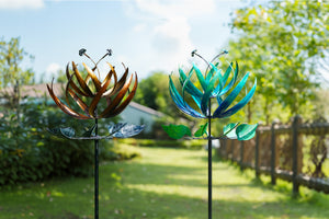 This is a garden scene featuring a decorative wind spinner. The wind spinner has a beautiful, multi - layered, petal - like structure in shades of blue and green. It's mounted on a black pole, with green leaf - shaped elements near the bottom. To the left, there are white flowers and green foliage in front of a light - colored house with a window. On the right, there's a spiky green plant. In the background, a wooden fence and a clear blue sky complete the setting, creating a pleasant and vibrant garden atm
