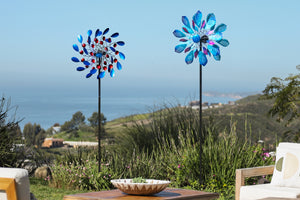 This image shows two windmills spinning in the wind amidst a vast natural landscape. The azure sea and rolling mountains in the distance create a tranquil and open atmosphere. The vibrant red and blue colors of the windmills, dotted among the lush green grass, add a touch of liveliness and vitality to this tranquil scene.