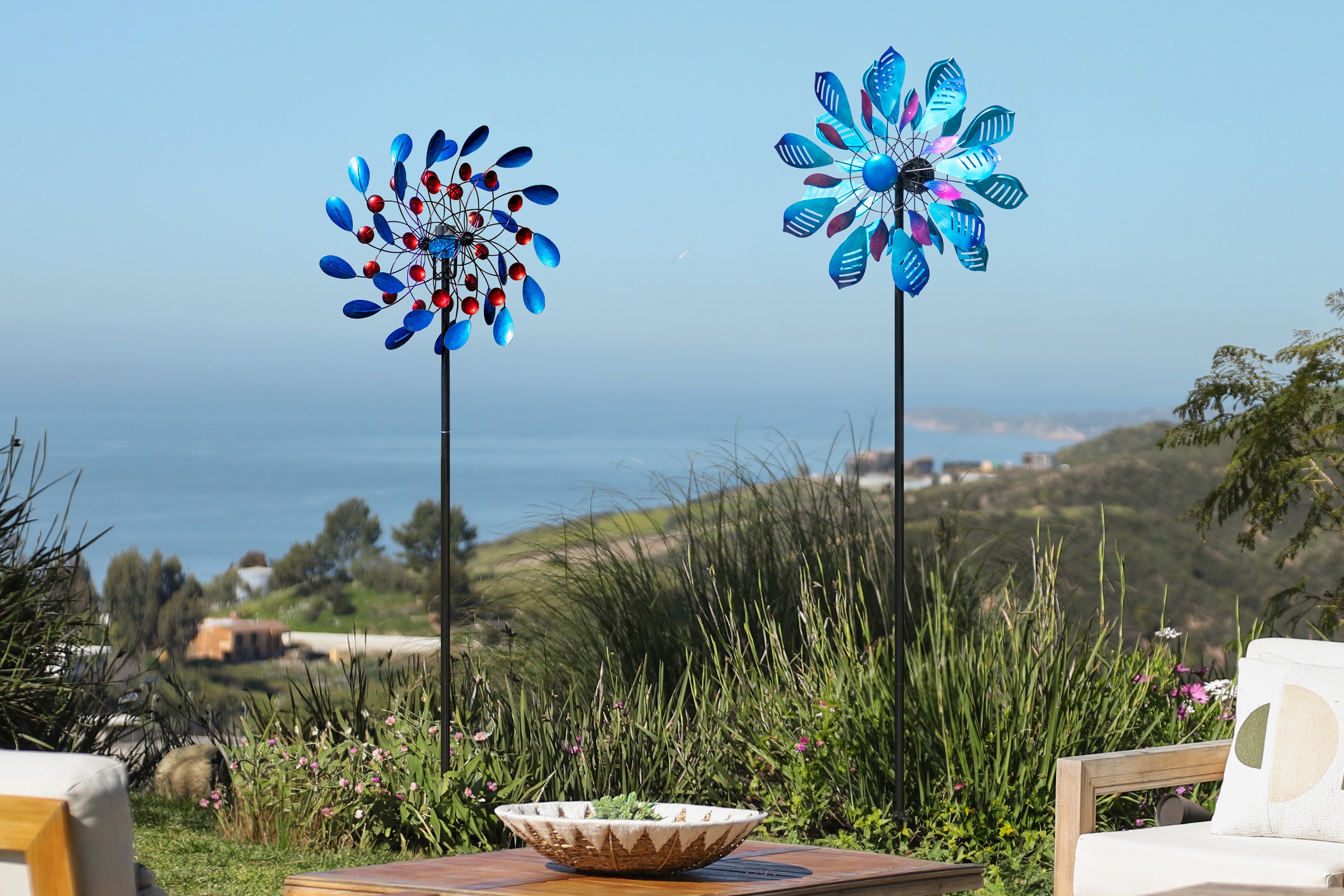 This image shows two windmills spinning in the wind amidst a vast natural landscape. The azure sea and rolling mountains in the distance create a tranquil and open atmosphere. The vibrant red and blue colors of the windmills, dotted among the lush green grass, add a touch of liveliness and vitality to this tranquil scene.