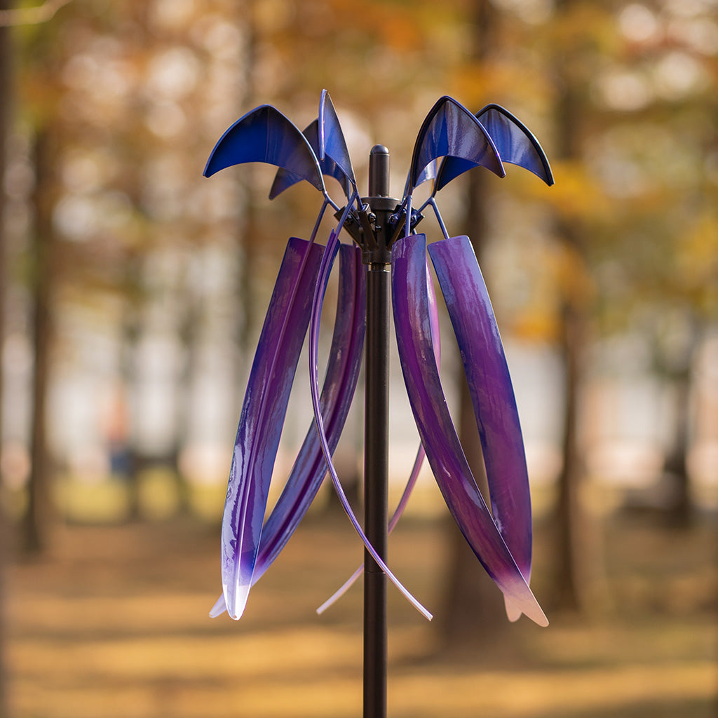 A metal garden wind spinner with purple and blue bat-wing shaped blades on a black pole, photographed outdoors against a blurred autumn park background.