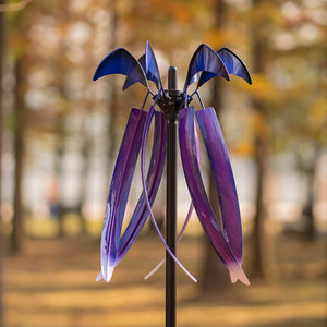 A metal garden wind spinner with purple and blue bat-wing shaped blades on a black pole, photographed outdoors against a blurred autumn park background.