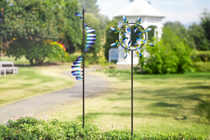 This image shows two windmills, each standing beside a beautiful garden path. The left windmill's spiral blades, with a blue gradient design, create a dynamic effect. The right windmill, with its circular structure and evenly spaced, colorful blades, resembles a blooming flower. In the background, a white pavilion and surrounding greenery create a tranquil and pleasant scene.